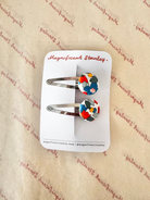 A pair of hair clips with colorful geometric mosaic designs on silver metal bars, displayed on a white card labeled 'Magnificent Stanley' against a beige patterned background.