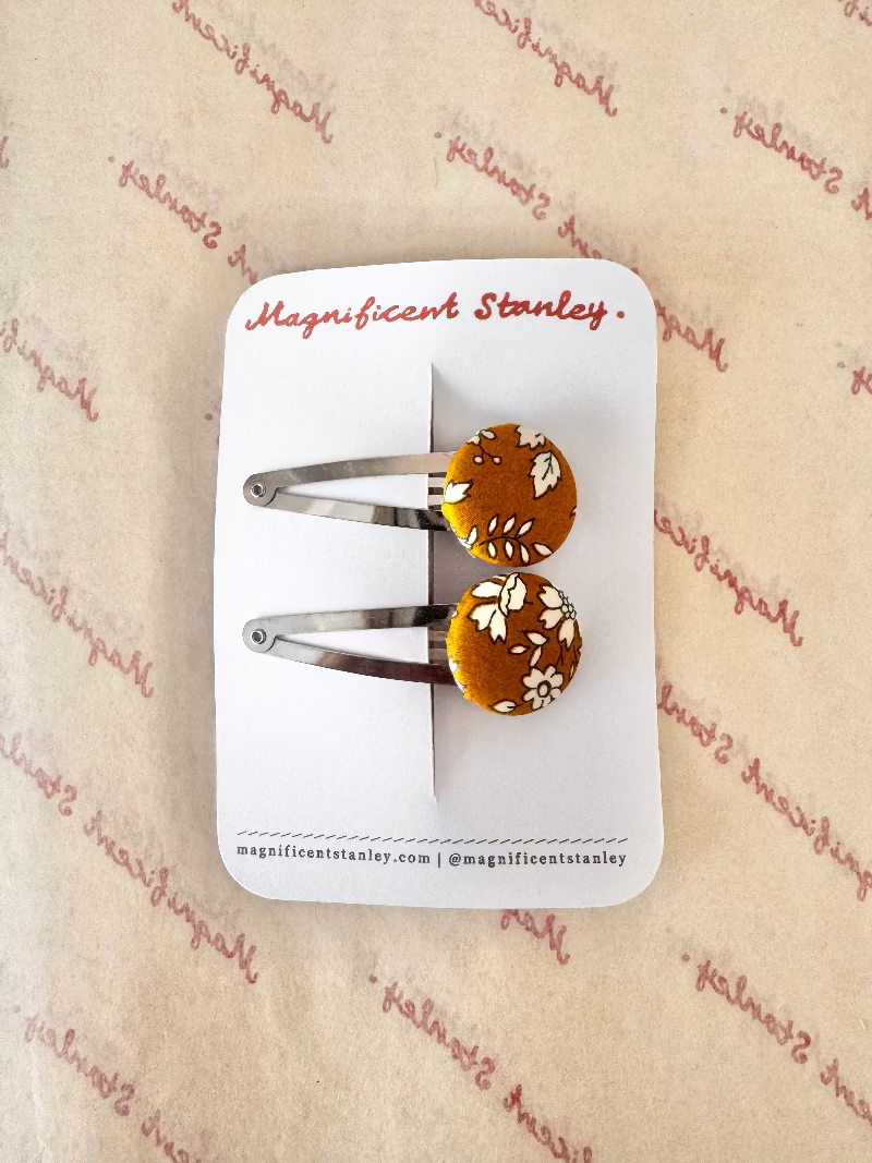 Two small hair clips with round orange-brown floral buttons and silver metal bars, attached to a white display card labeled 'Magnificent Stanley' and placed on patterned paper.