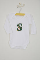 A white long-sleeve baby bodysuit laid flat on a white surface, featuring a decorative capital letter 'S' on the chest with a green Liberty Elves pattern.