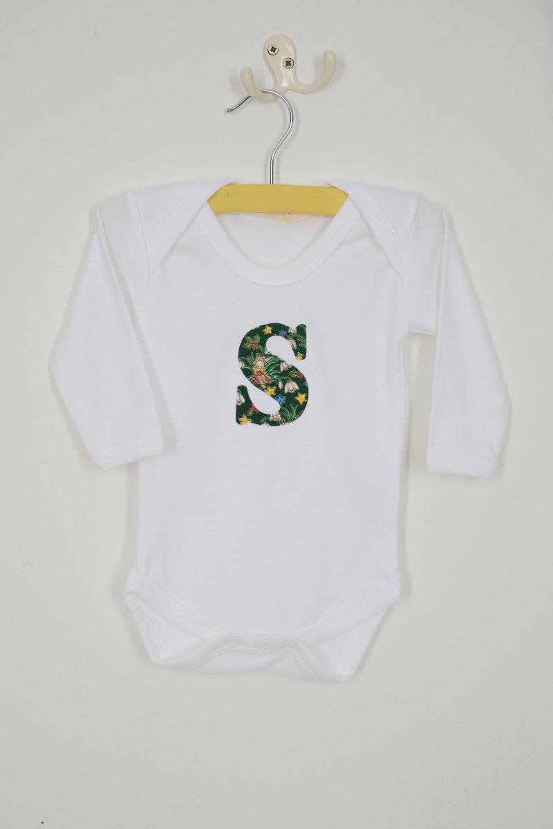 A white long-sleeve baby bodysuit laid flat on a white surface, featuring a decorative capital letter 'S' on the chest with a green Liberty Elves pattern.