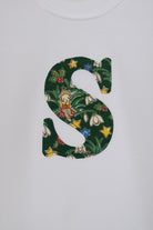 A close up of a white long-sleeve baby bodysuit laid flat on a white surface, featuring a decorative capital letter 'S' on the chest with a green Liberty Elves pattern.