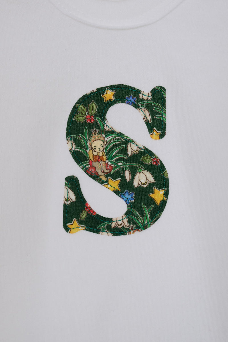 A close up of a white long-sleeve baby bodysuit laid flat on a white surface, featuring a decorative capital letter 'S' on the chest with a green Liberty Elves pattern.