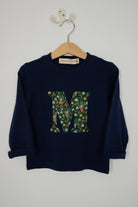 A navy blue long-sleeve cotton t-shirt with a large letter 'M' graphic on the chest filled with a colourful toadstool, snowdrop, holly and elf Liberty pattern, hanging on a wooden hanger against a light grey wall.