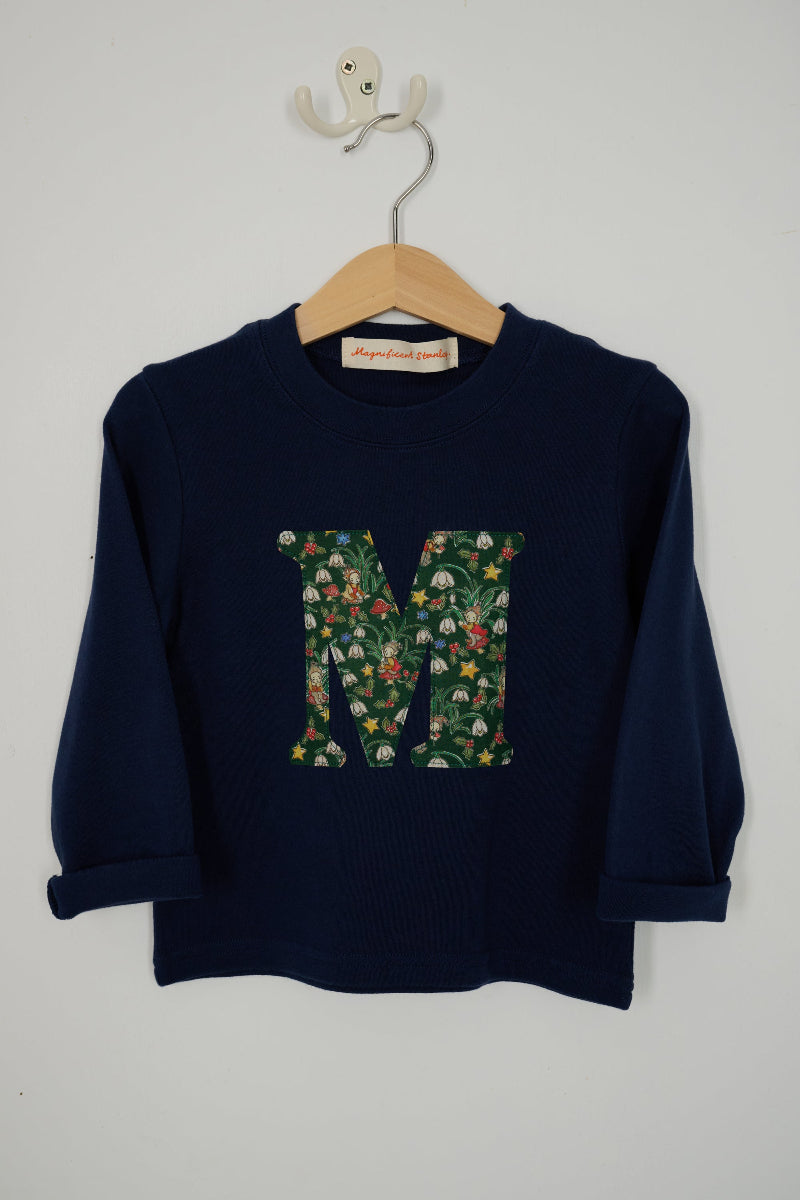 A navy blue long-sleeve cotton t-shirt with a large letter 'M' graphic on the chest filled with a colourful toadstool, snowdrop, holly and elf Liberty pattern, hanging on a wooden hanger against a light grey wall.