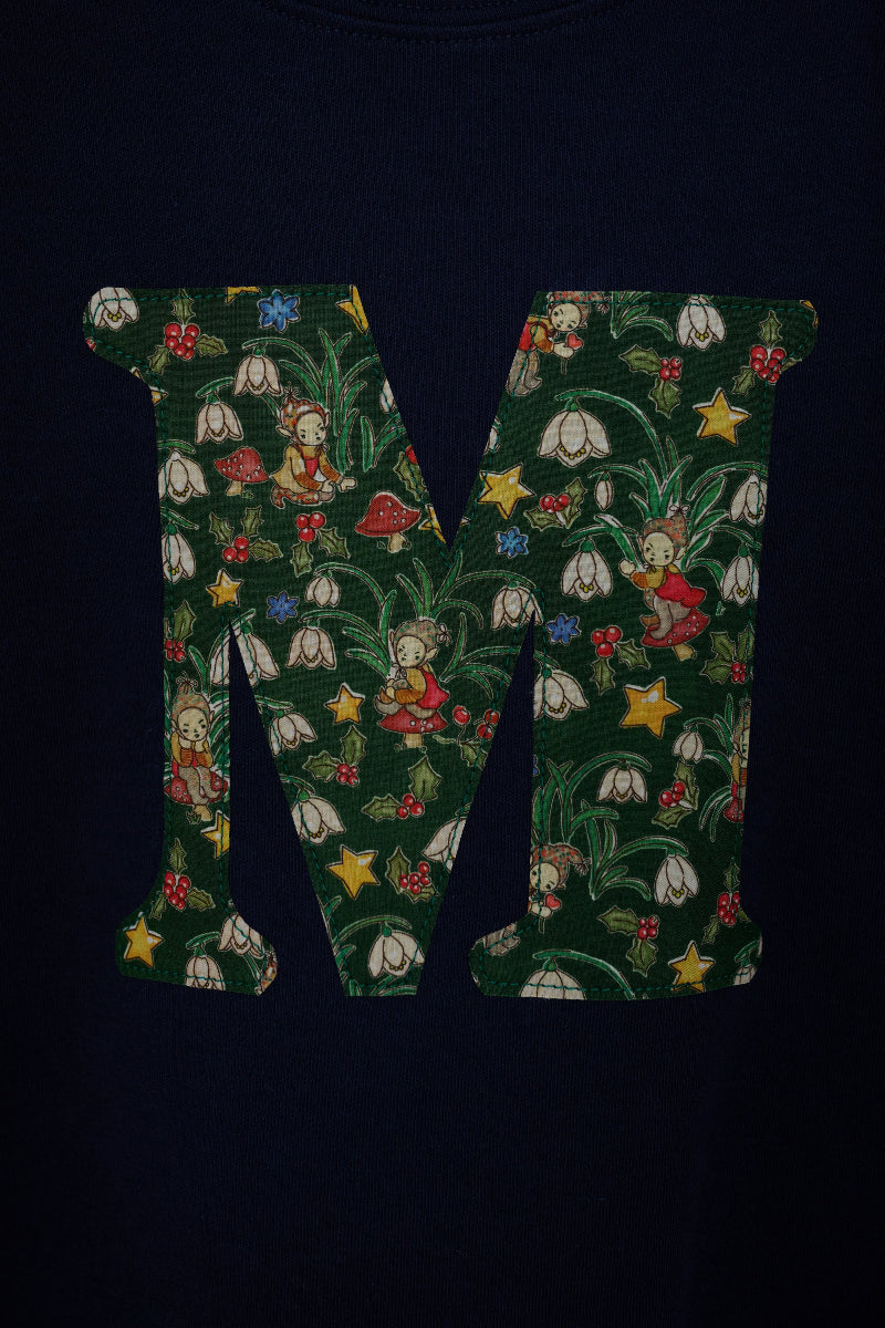 A close-up of a dark navy blue cotton kids t-shirt featuring a large letter 'M' printed on the front, filled with a detailed Liberty woodland fabric of green leaves, white snowdrop flowers, red mushrooms, elves and yellow stars.