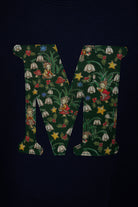 A close-up of a dark navy blue cotton kids t-shirt featuring a large letter 'M' printed on the front, filled with a detailed Liberty woodland fabric of green leaves, white snowdrop flowers, red mushrooms, elves and yellow stars.