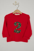 A red long-sleeve children's shirt hanging on a wooden hanger with a white wall hook behind it. The shirt features a large number '2' in Liberty Elves print .