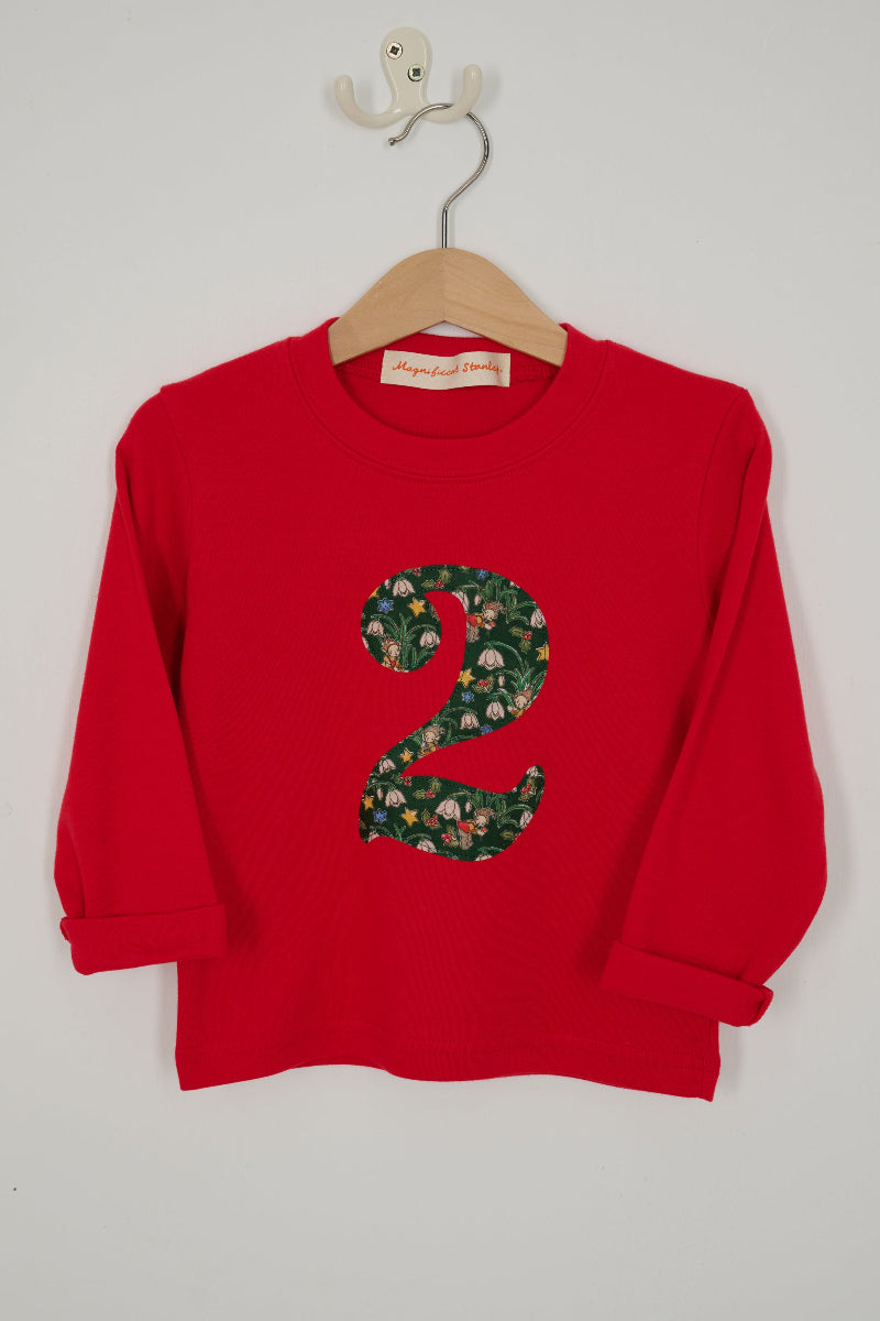 A red long-sleeve children's shirt hanging on a wooden hanger with a white wall hook behind it. The shirt features a large number '2' in Liberty Elves print .