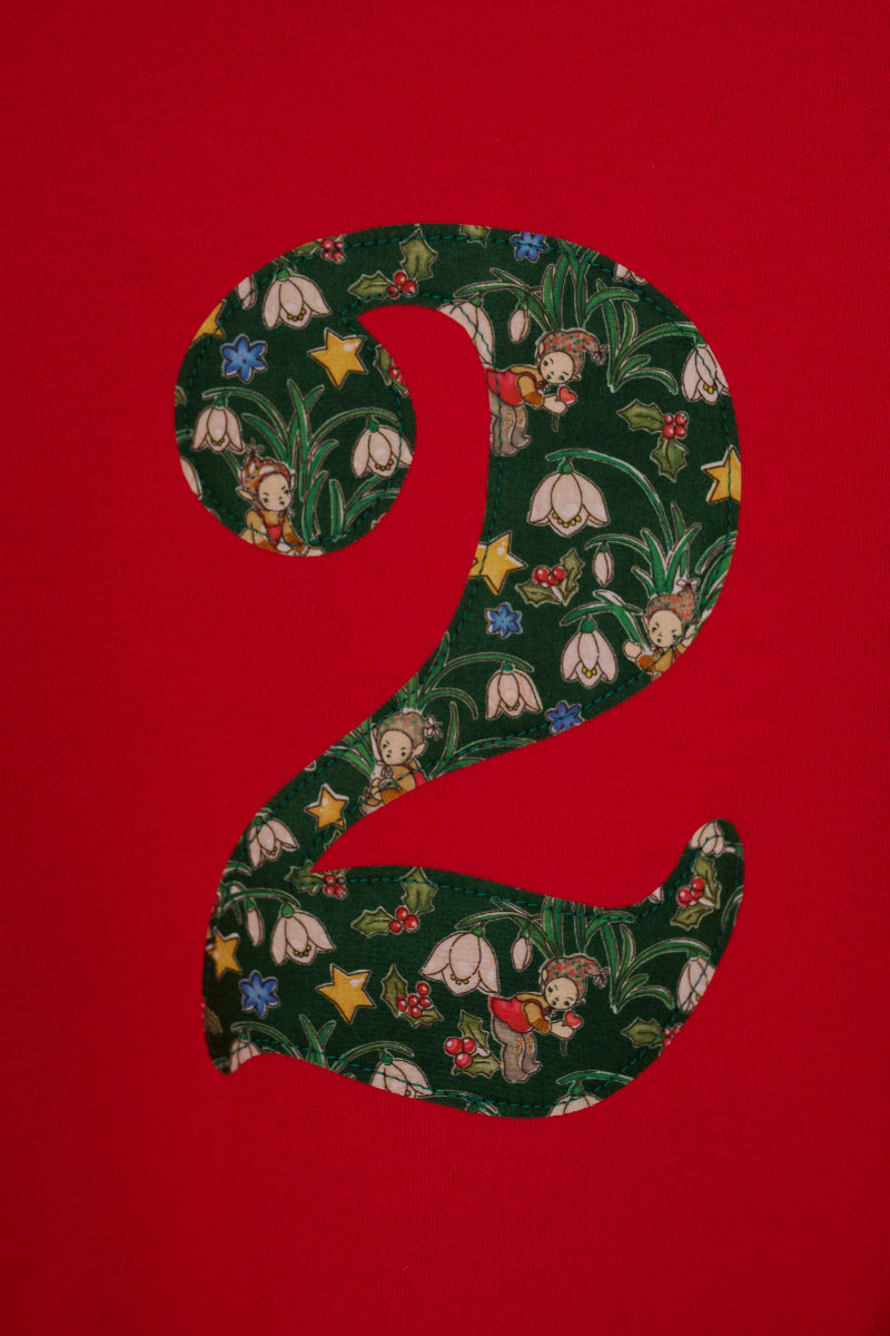 A large Liberty fabric number 2 filled with a dark green fabric pattern featuring white snowdrops, small fairy elves, and yellow stars, set against a solid bright red background.