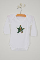 A white long-sleeve christmas baby grow with a green star-shaped patch featuring a festive Liberty Elves print featuring snowdrops, holly, elves and stars. 