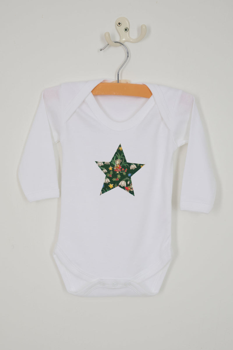 A white long-sleeve christmas baby grow with a green star-shaped patch featuring a festive Liberty Elves print featuring snowdrops, holly, elves and stars. 