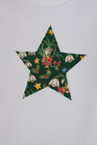 A white long-sleeve baby bodysuit with a green star-shaped patch featuring a festive Liberty Elves print featuring snowdrops, holly, elves and stars, laid flat on a green patterned fabric background. A close up of the appliqué stitching 