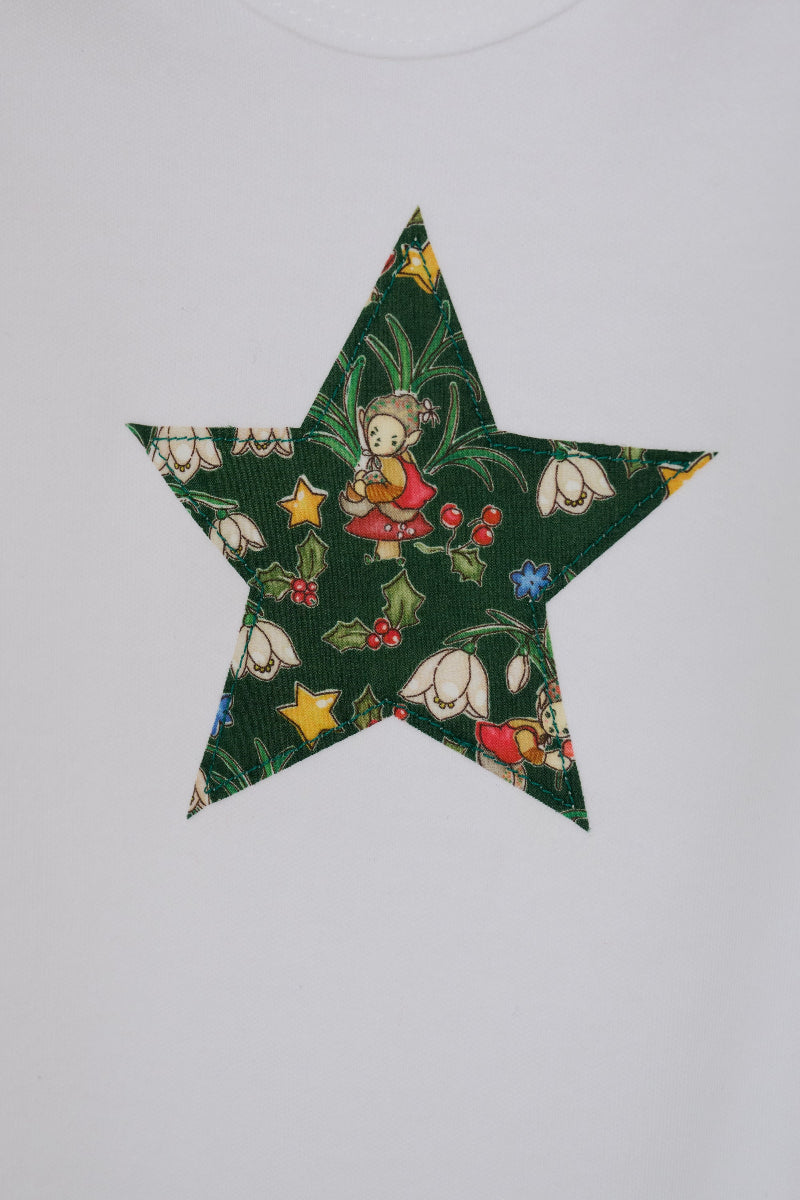 A white long-sleeve baby bodysuit with a green star-shaped patch featuring a festive Liberty Elves print featuring snowdrops, holly, elves and stars, laid flat on a green patterned fabric background. A close up of the appliqué stitching 