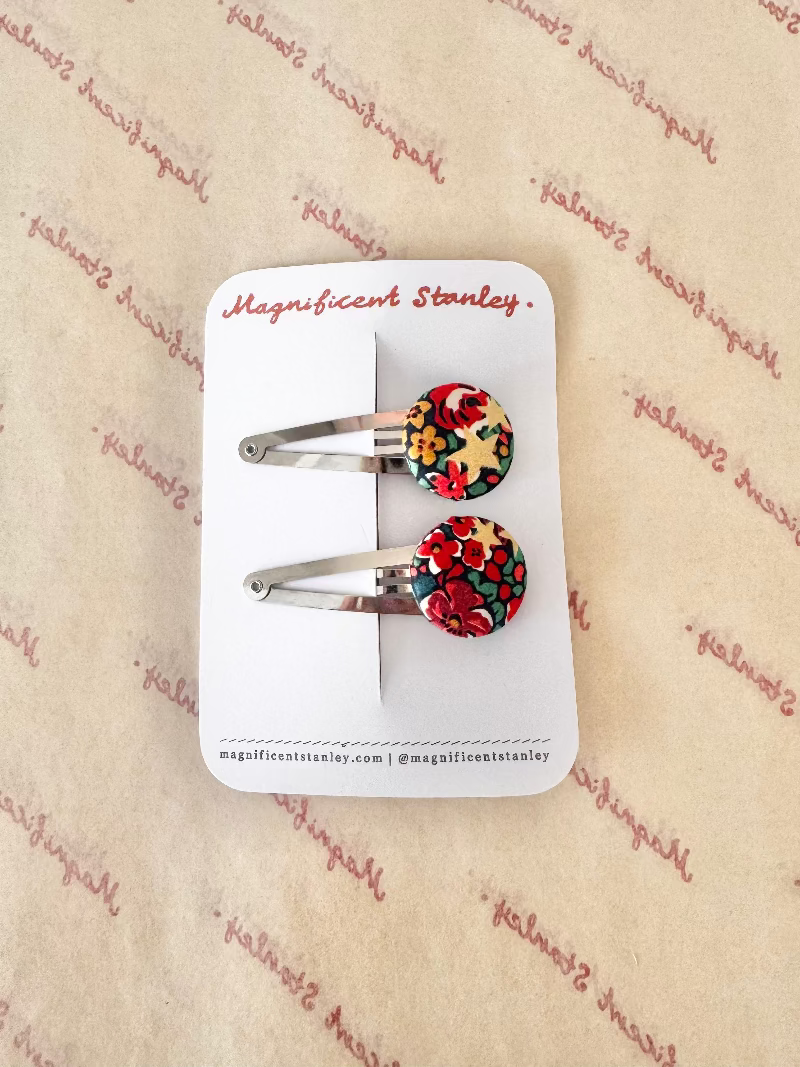 A pair of silver hair clips with round floral cabochons in red, yellow, and green, mounted on a white display card branded 'Magnificent Stanley' against a beige patterned background.