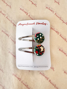 Two small barrette hair clips with round decorative tops on a white display card. The clips have silver metal bars and round cabochons featuring a green background with red, white, and yellow star-and-stripe patterns.