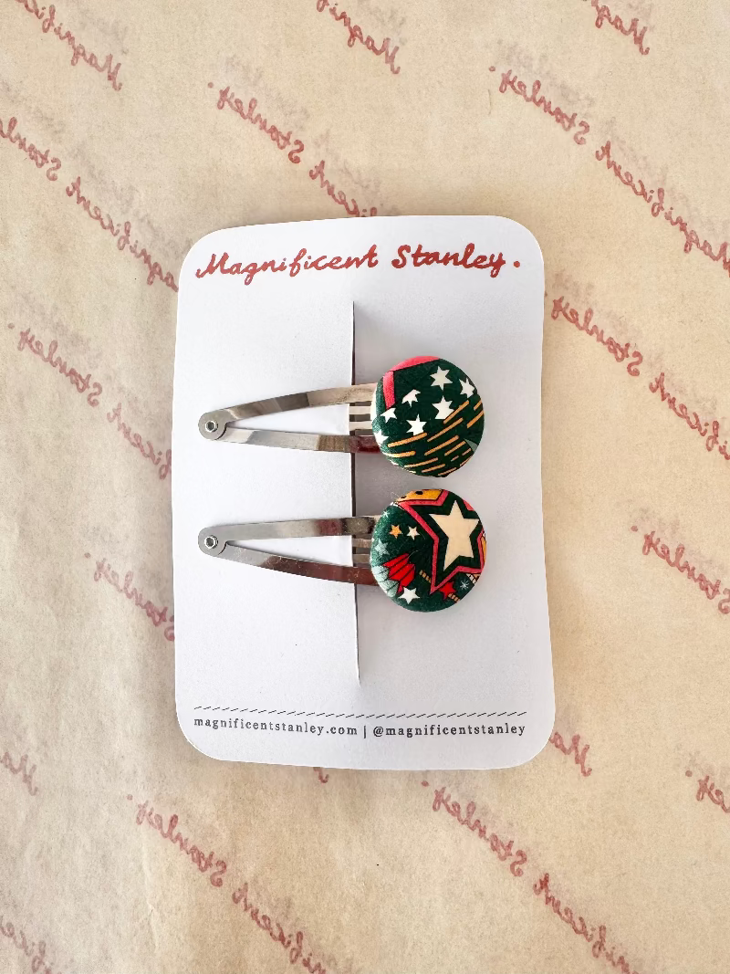 Two small barrette hair clips with round decorative tops on a white display card. The clips have silver metal bars and round cabochons featuring a green background with red, white, and yellow star-and-stripe patterns.