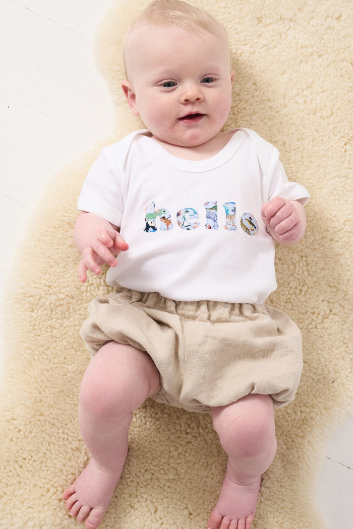 A 3 month old baby wearing a white cotton babygro with the word hello sewn on the front, cut from Quey 2 zoo animal  Liberty print.