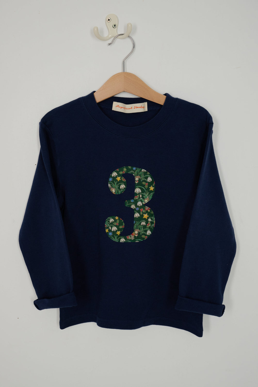 A navy blue long-sleeve kids t-shirt hanging on a wooden hanger against a plain light gray wall. The front features a large number '3' graphic filled with a green floral pattern called Liberty Elves.