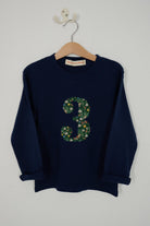 A navy blue long-sleeve kids t-shirt hanging on a wooden hanger against a plain light gray wall. The front features a large number '3' graphic filled with a green floral pattern called Liberty Elves.