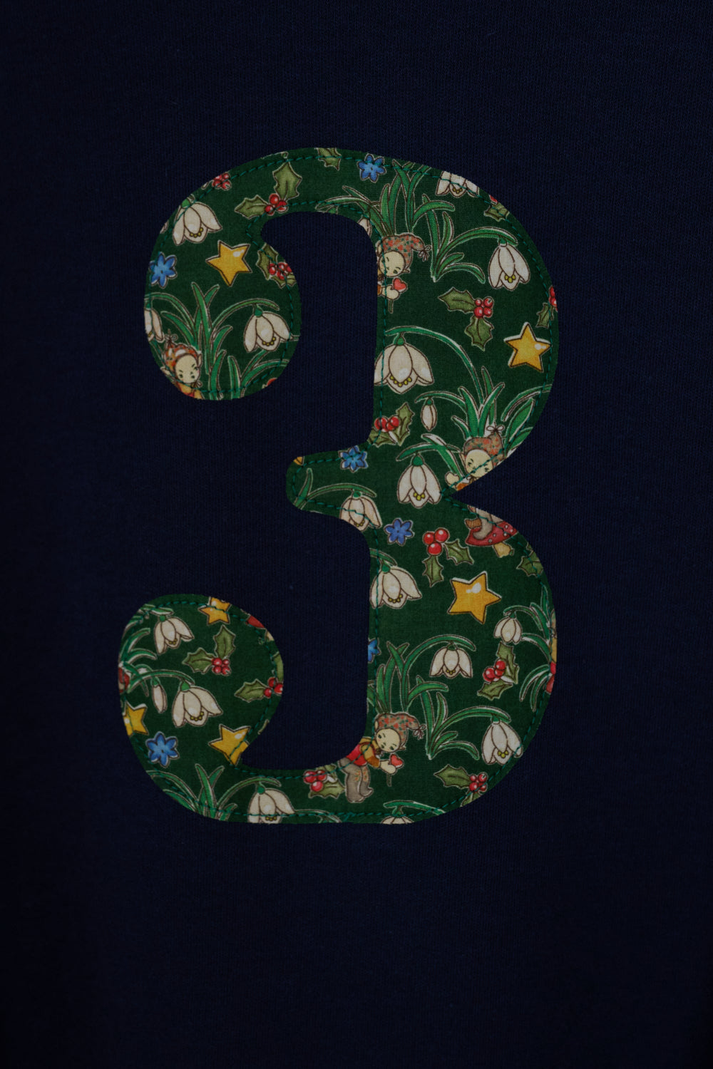 A navy blue kids cotton t-shirt laid flat on  featuring a large number '3' on the front filled with a green holiday-themed pattern of pinecones, ornaments, and snowdrops called Liberty Elves. A close up of the applique sewing detail.