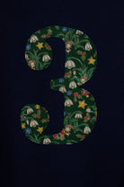 A navy blue kids cotton t-shirt laid flat on  featuring a large number '3' on the front filled with a green holiday-themed pattern of pinecones, ornaments, and snowdrops called Liberty Elves. A close up of the applique sewing detail.