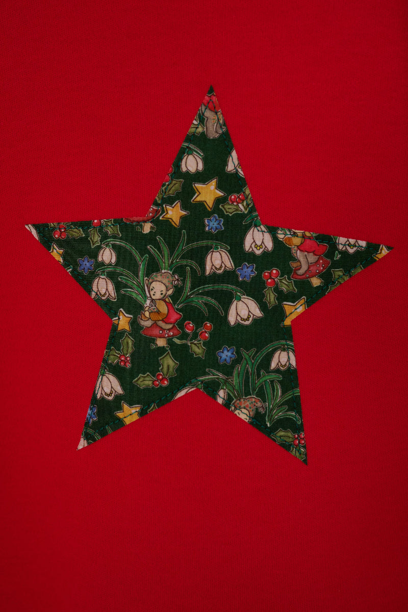 A red long-sleeve sweatshirt with a large star-shaped patch on the chest featuring a Christmas Liberty Elves print. The print features snowdrops, holly, elves, toadstools and stars.