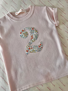 A light pink short-sleeve children's T-shirt laid flat on a patterned cream surface, featuring a large floral number 2 applique on the front.
