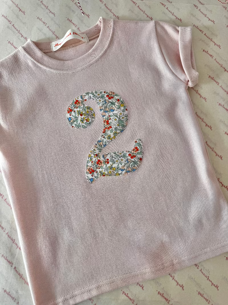 A light pink short-sleeve children's T-shirt laid flat on a patterned cream surface, featuring a large floral number 2 applique on the front.