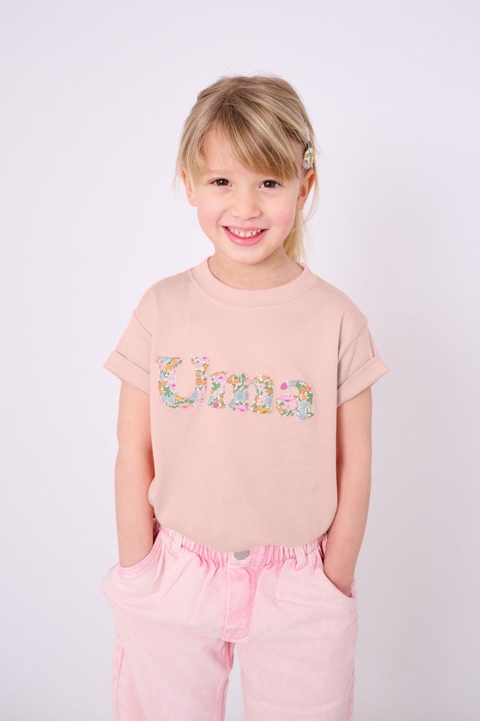 A 4 year old girl wearing a soft pink kids t-shirt with her name cut from Liberty print in lowercase letters.