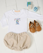 A baby outfit flat lay featuring a white long-sleeve bodysuit with a colorful letter 'T' in Liberty Quey 2 print, paired with beige bloomers and tan lace-up shoes, styled with wooden blocks on a white wood plank background.