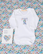 A white long-sleeve baby bodysuit laid flat on a colourful animal-print  Quey 2 Liberty fabric background, with a large letter T printed on the chest in a pastel multicolour pattern. A small white card reading 'hello baby!' is placed beside it.
