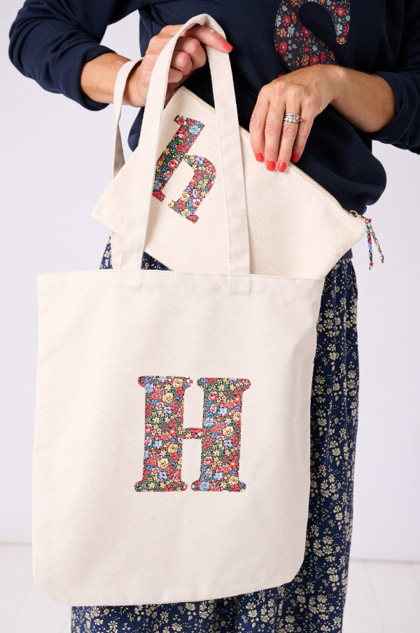 A woman with a Canvas Tote Bag on her shoulder, personalised with her initial in Liberty ditsy floral fabric. She is taking out a matching personalised Liberty print wash bag