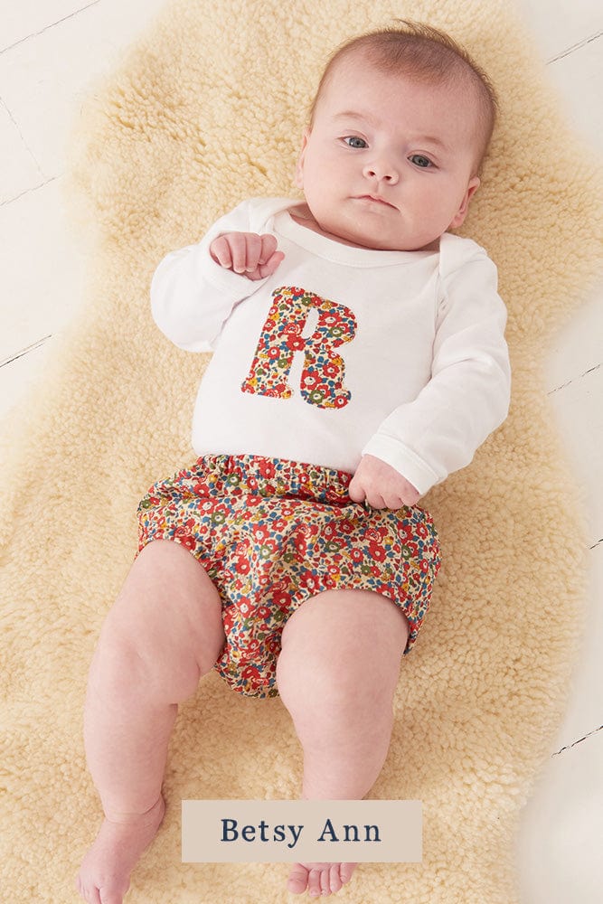 A smiling newborn baby wearing a personalised white bodysuit with an initial on the front cut from Liberty floral fabric.