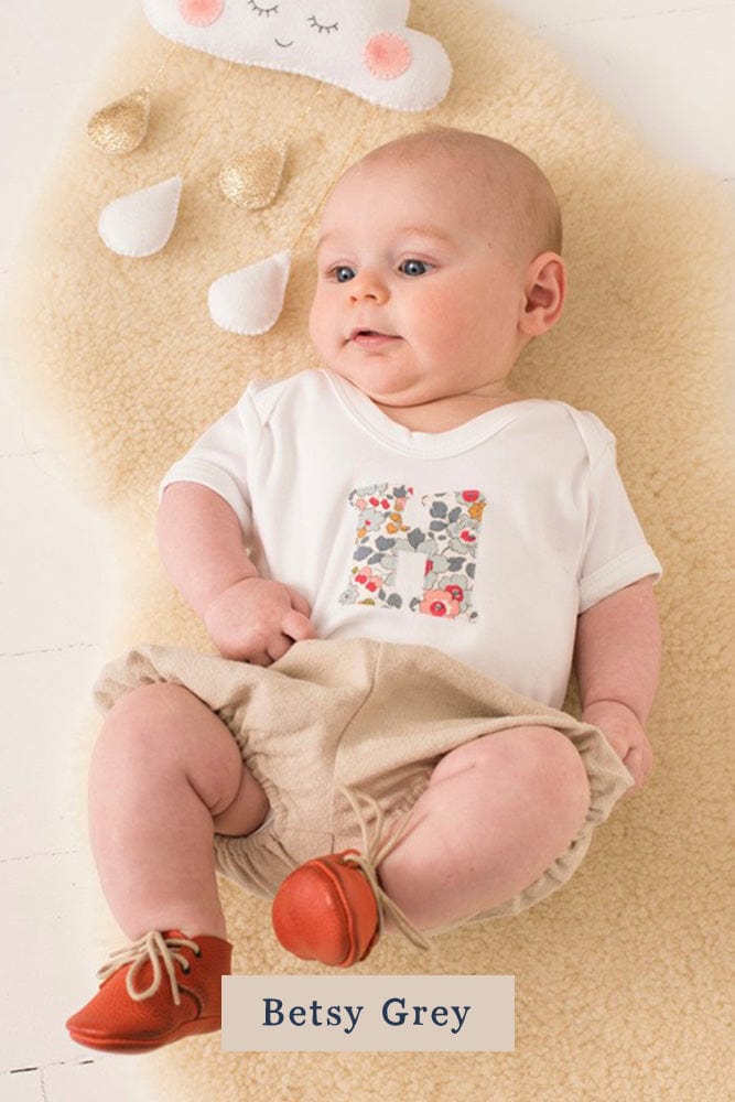 A smiling newborn baby wearing a personalised white bodysuit with an initial on the front cut from betsy grey Liberty floral fabric.