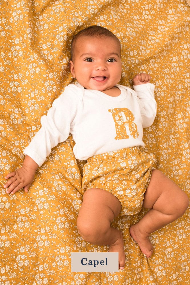 A smiling newborn baby wearing a personalised white bodysuit with an initial on the front cut from Capel Liberty floral fabric.