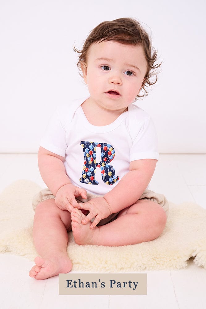 A smiling newborn baby wearing a personalised white bodysuit with an initial on the front cut from Ethan's party balloon Liberty balloon fabric.