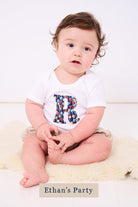 A smiling newborn baby wearing a personalised white bodysuit with an initial on the front cut from Ethan's party balloon Liberty balloon fabric.