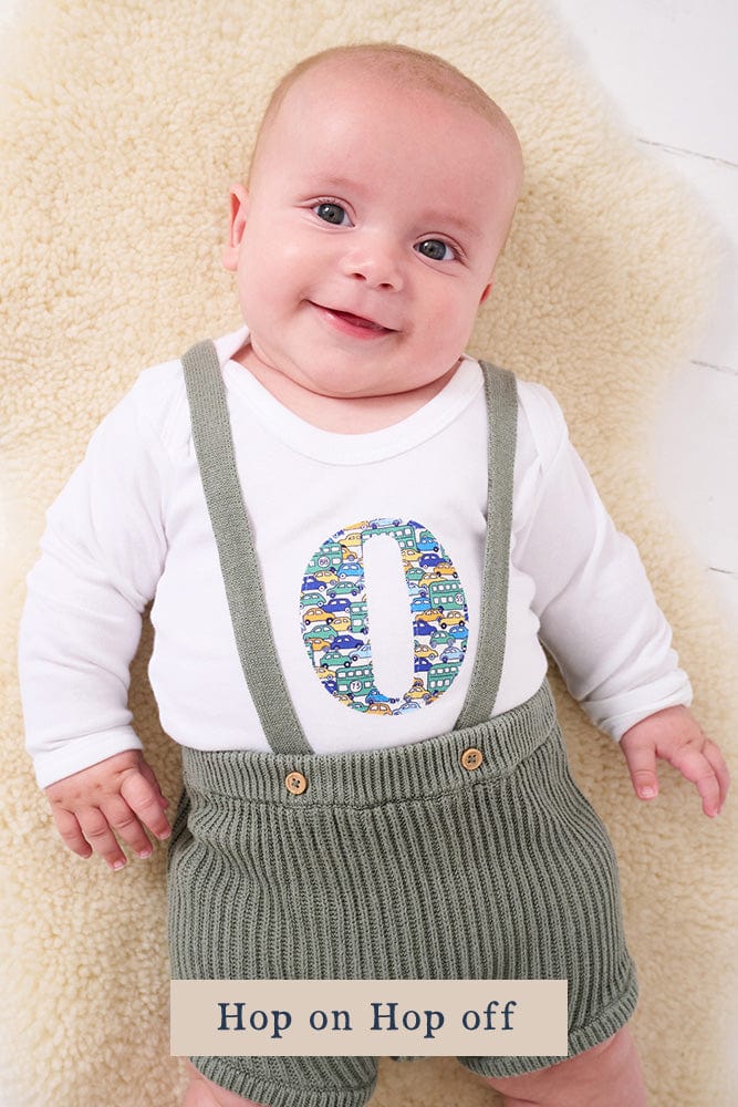 A smiling newborn baby wearing a personalised white bodysuit with an initial on the front cut from Liberty cars fabric.
