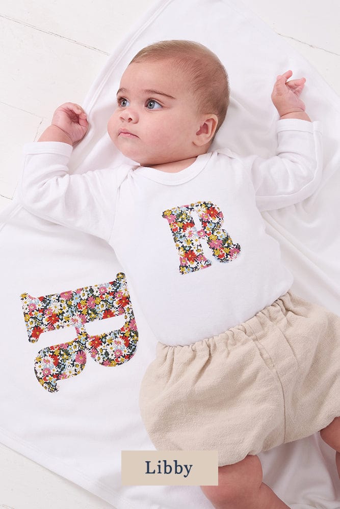 A smiling newborn baby wearing a personalised white bodysuit with an initial on the front cut from Liberty floral fabric. She is laying on a matching personalised blanket.