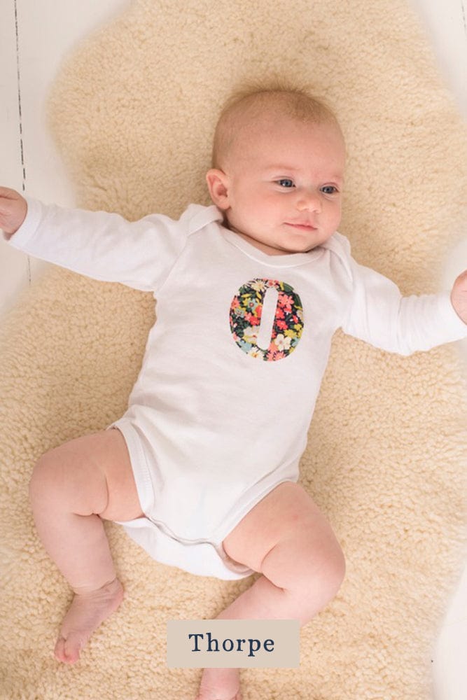 A smiling newborn baby wearing a personalised white bodysuit with an initial on the front cut from Liberty floral fabric.