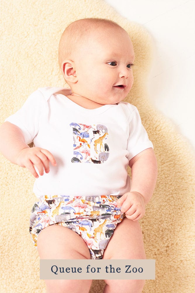 A smiling newborn baby wearing a personalised white bodysuit with an initial on the front cut from Liberty zoo animal fabric.