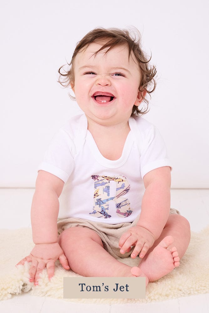 A smiling newborn baby wearing a personalised white bodysuit with an initial on the front cut from Liberty jet plane fabric.