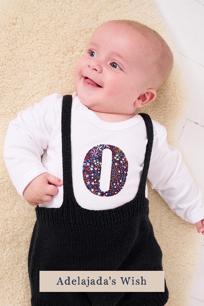 A smiling newborn baby wearing a personalised white bodysuit with an initial on the front cut from Liberty Adelajda starry fabric.