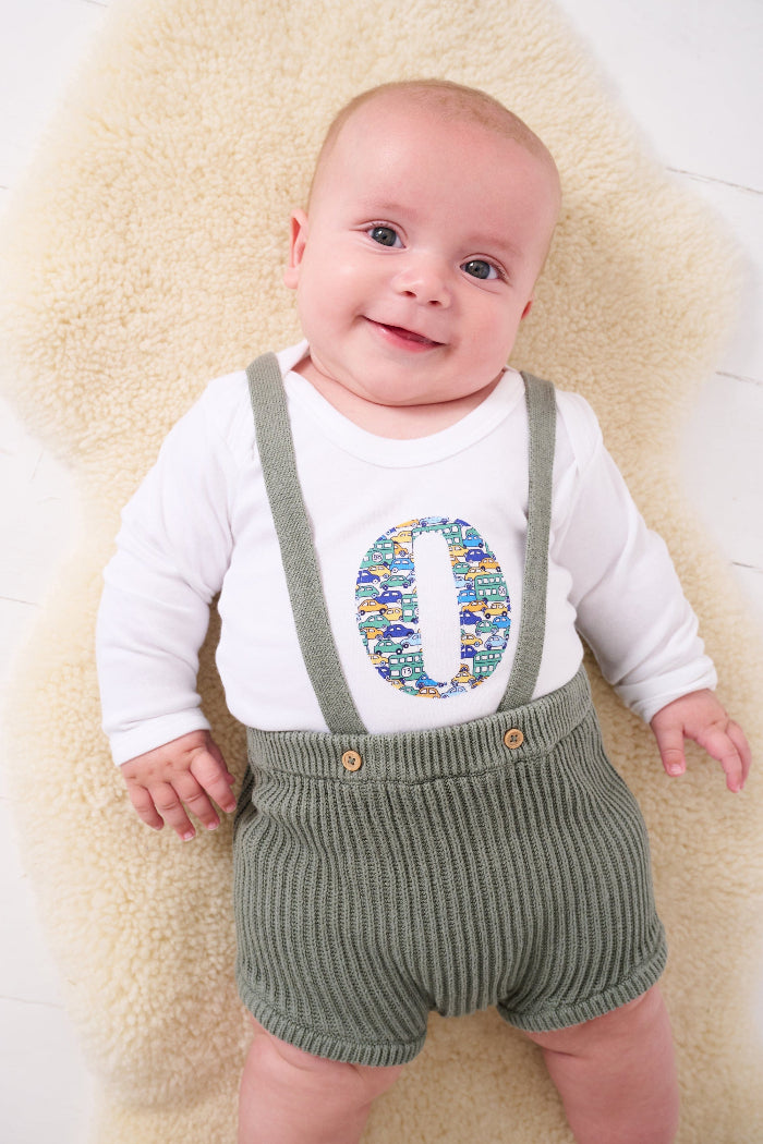A baby wearing a Personalised Bodysuit in hop on hop off Liberty Print featuring vintage cars and busses.