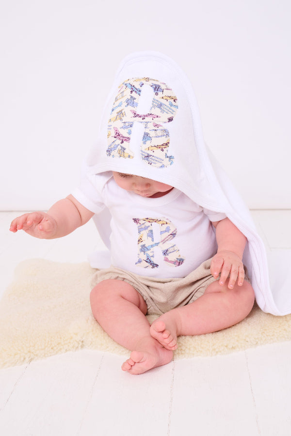 A 6 month old baby boy wearing a Personalised Bodysuit in Tom's Jet Liberty Print with his initial sewn onto the front. He is also wearing a personalised hooded towel.