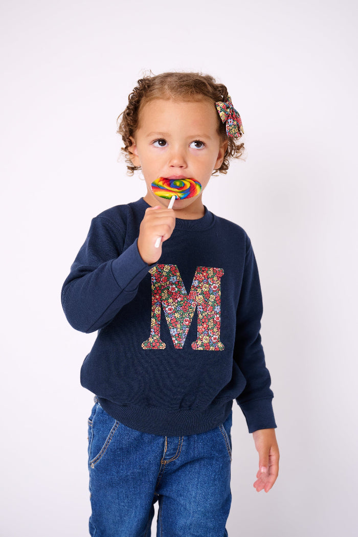 A 3 year old girl wearing a  navy Magnificent Stanley personalised Navy Sweatshirt with her initial on the front in floral emma louise liberty print.