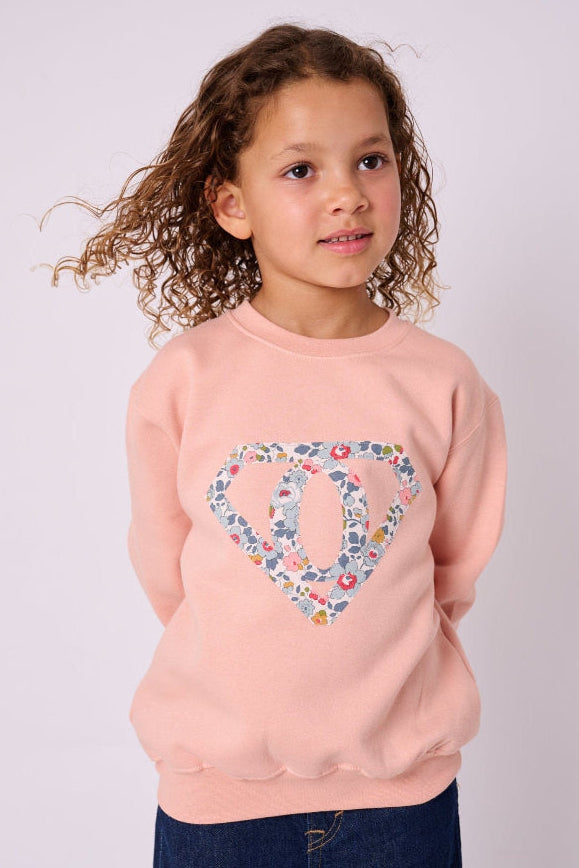 A young girl wearing a dusty pink sweatshirt with her initial sewn on the front in a Superhero motif made from floral Liberty Print.