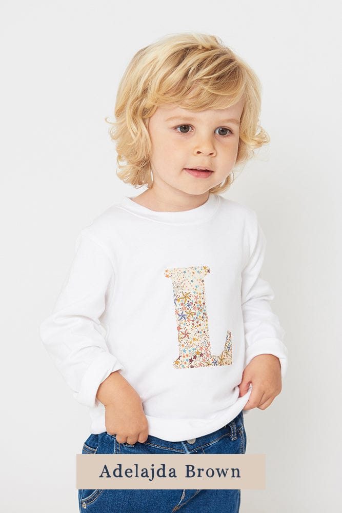 A 3 year old boy wearing a personalised white cotton t-shirt with her initial on in Adelajda star Liberty fabric.