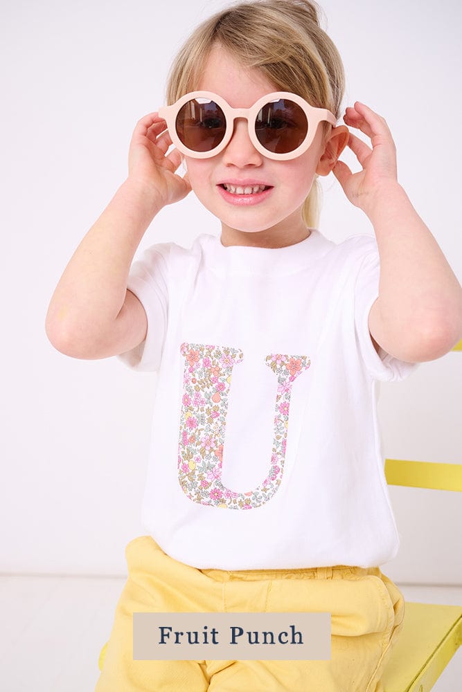 A 3 year old girl wearing a personalised white cotton t-shirt with her initial on in Fruit Punch floral Liberty fabric.