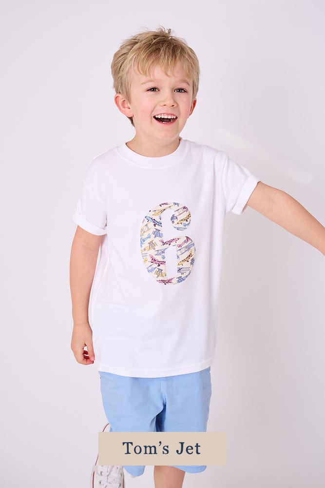 A 6 year old boy wearing a birthday white cotton t-shirt with the number 6 on in jet planes Liberty fabric.